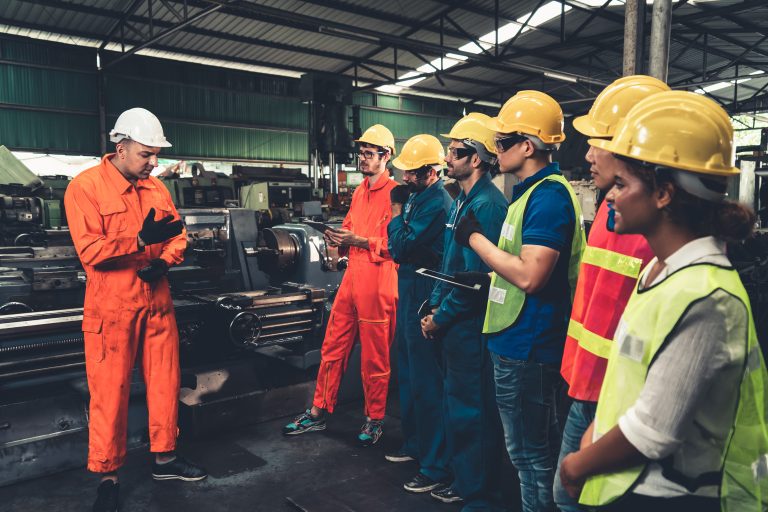 A group of workers in a factory setting, wearing safety gear including hard hats, reflective vests, and gloves. They are attentively listening to a supervisor in an orange jumpsuit who is providing safety instructions. The background shows industrial equipment and machinery, highlighting a focus on workplace safety. Focusing on workplace safety is a best practice for a strong Workers Compensation Insurance program.